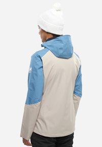 Wasserabweisende Jacke mit einem beigen Oberteil und blauen Ärmeln sowie Kapuze. Tailliertes Design mit strukturiertem Finish und Logodetail am Ärmel.