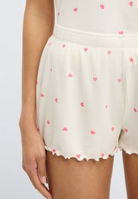 Ελαφριά κρεμ σορτς με ραβδώσεις κα ι διασκορπισμένα ροζ μοτίβα σε σχήμα καρδιών. Στρογγυλεμένο τελείωμα με βολάν. Ελαστική ζώνη στη μέση.