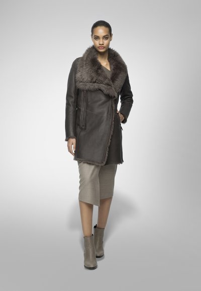Cappotto di pelle marrone con colletto in pelliccia sintetica, dotato di una tasca con zip. Indossato su una gonna grigio chiara a ginocchio e stivaletti beige.