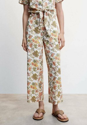 Femme portant un pantalon large à fleurs avec un motif de feuilles d'automne, taille nouée et sandales ouvertes couleur tan, debout sur un sol en béton.