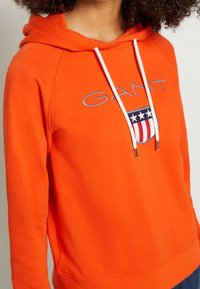 Pomarańczowa bawełniana bluza z kapturem z kieszenią kangurką, białymi sznurkami oraz ozdobnym logo z gwiazdami i paskami na piersi.