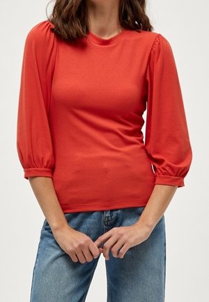 Kvinde iført en tætsiddende rød top med puffede trekvart ærmer og blå denimjeans, stående mod en ensfarvet baggrund.