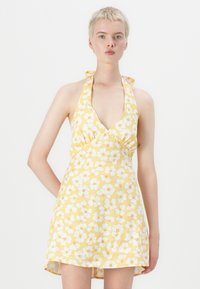 Florale gele halterjurk met een diepe V-hals, versierd met witte bloemen en oranje accenten op een gestructureerde stof. Korte, uitlopende zoom.