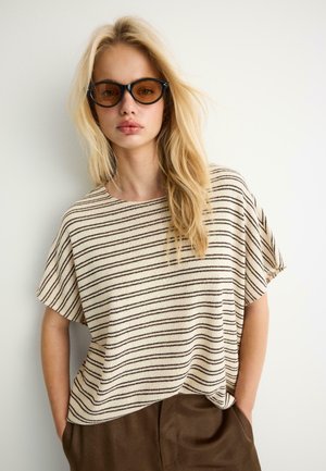 Plavovláska s hnedými tónovanými slnečnými okuliarmi, voľnou béžovou pruhovanou košeľou a hnedými nohavicami, stojaca pred jednoduchou bielou stenou.