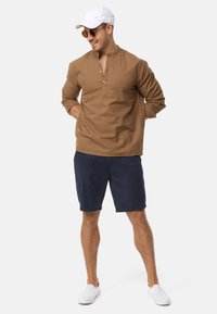 Brun langærmet skjorte med krave og knaplukning, parret med navy shorts og hvide slip-on sko; solbriller og en hvid kasket inkluderet.
