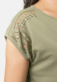 Camiseta de manga corta en verde oliva con decoración de cuentas y bordados en naranja y plata en el hombro y la parte superior del brazo.