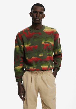 Green camo-patterned sweater with red and yellow accents, made of textured knit fabric. Long sleeves, crew neckline, worn with beige pants.