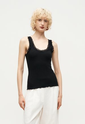 Femme aux cheveux blonds courts et bouclés, portant un haut sans manches noir orné de dentelle et un pantalon blanc ample, debout devant un fond uni.