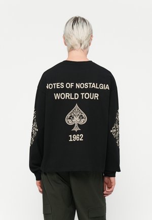 Black long-sleeve sweatshirt with "NOTES OF NOSTALGIA WORLD TOUR 1962" printed in cream on the back and decorative designs on the sleeves.