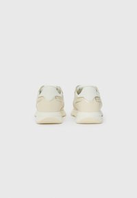 Sneakers di un crema chiaro con una parte superiore testurizzata, tallone liscio e sottili accenti dorati. Forma arrotondata con suola ammortizzata per il comfort.
