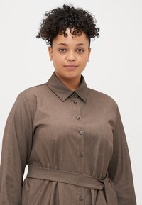 Barna gombos ruházat gallérral, hosszú ujjakkal és megkötött derékkal. Texturált anyagból, finom fényű, és hozzáillő gombokkal készült.