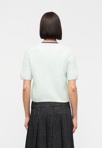 Γυναίκα με καστανά, ίσια, μαλλιά μέχρι τους ώμους, που φοράει ανοιχτό μπλε πουλόβερ με κοντά μανίκια και σκούρα πλισέ φούστα, με το πρόσωπο στραμμένο προς τα πίσω.