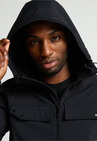 Melnā kapuce ar rāvējslēdzēju un divām priekšējām kabatām. Gluda, struktūra auduma ar redzamām šuvēm un regulējamām kapuces auklām.