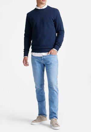 Man die een marineblauwe trui draagt over een wit overhemd, lichtblauwe spijkerbroek en beige sneakers, staand met één hand in de zak.