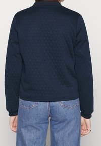 Granatowa pikowana kurtka z okrągłym kołnierzem, ściągaczami na rękawach i skróconym dołem, zestawiona z jasnoniebieskimi jeansami o prostych nogawkach.