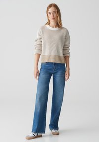 Hellbeige und weiße gestreifte Strickpullover mit gerippter Textur, kombiniert mit hellblauen weiten Jeans. Das Model trägt weiße Sneaker mit grauen Akzenten.