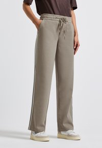 Persoon in taupekleurige wijde sweatpants met witte en zwarte zijkanten, gecombineerd met witte sneakers en een donkerbruin bovenstuk.