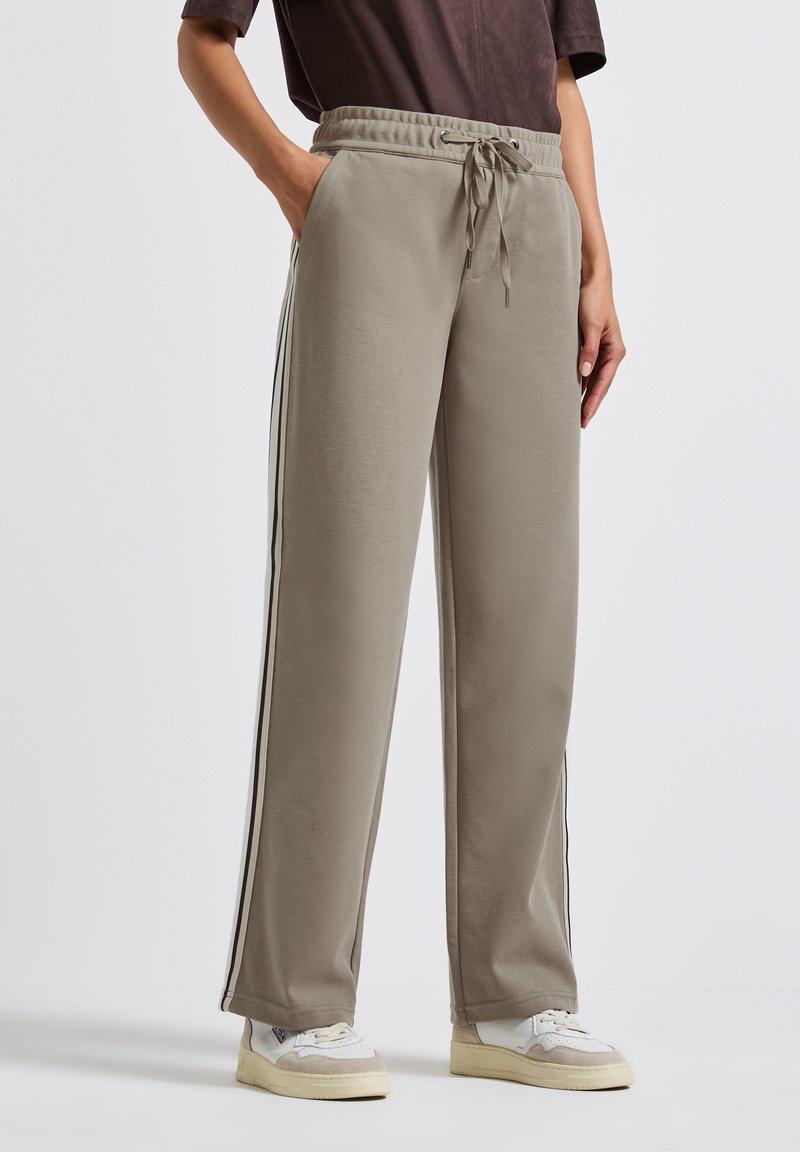 Persoon in taupekleurige wijde sweatpants met witte en zwarte zijkanten, gecombineerd met witte sneakers en een donkerbruin bovenstuk.