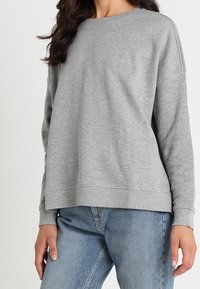 Grå sweatshirt med långa ärmar, rund halsringning och avslappnad passform. Tyget verkar mjukt med synliga sömnadsdetaljer. Bärs med blå jeans.