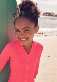 Roze geruite rashguard met een ritssluiting aan de voorkant, met lange mouwen en een nauwsluitend ontwerp, gedragen door een meisje dat bij een surfplank op het strand staat.