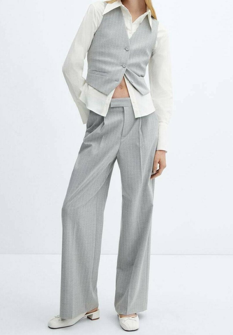 Femme portant un gilet tailored gris clair à fines rayures et un pantalon à jambes larges avec une chemise blanche boutonnée et des chaussures blanches à talons.