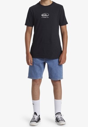 Person trägt ein schwarzes Quiksilver-T-Shirt, blaue Jeansshorts, weiße Sportsocken und schwarze niedrige Converse-Sneaker und steht aufrecht.
