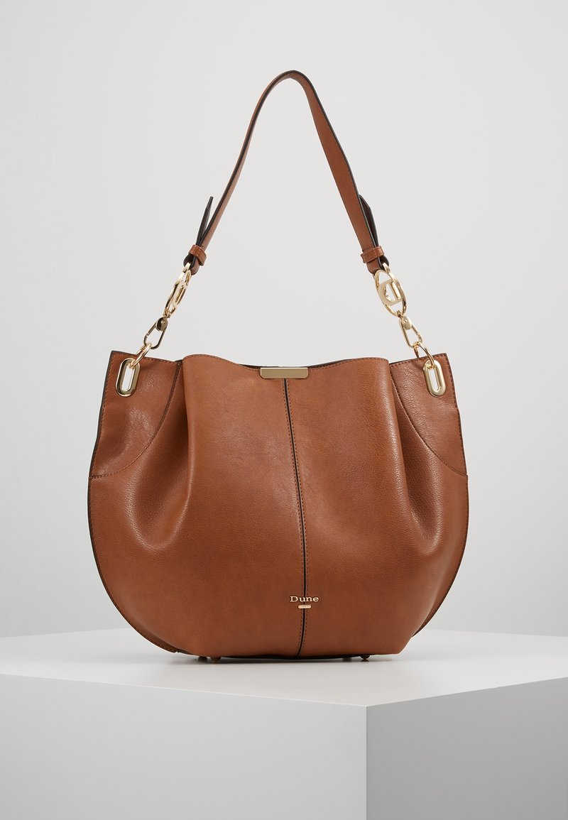 Sac à bandoulière en cuir marron avec ferrures dorées, une seule bandoulière et un petit logo "Dune" sur le devant, présenté sur une surface blanche.