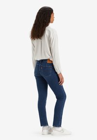 Žena s dugom kovrčavom kosom, odjevena u prugastu bijelu košulju, plave uske traperice i bijele tenisice, stoji naslonjena na jednoličnu pozadinu.