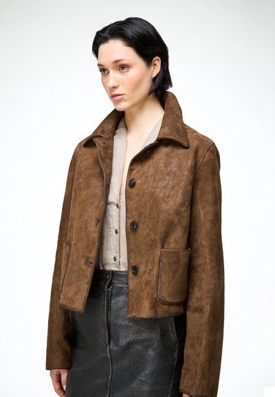 Femme aux cheveux courts et foncés portant une veste en daim marron boutonnée, un chemisier beige et une jupe noire texturée sur fond blanc.