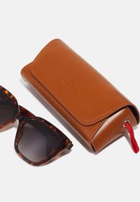 Occhiali da sole con montatura in tartaruga e lenti sfumate accanto a un astuccio in pelle marrone con una linguetta rossa e logo impresso.