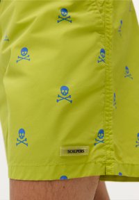 Shorts de bain vert lime avec un motif bleu de tête de mort et croix. Comprend une étiquette noire avec "SCALPERS" sur l'ourlet. Texture de tissu lisse.