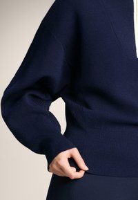 Marineblaue, gestrickte Pullover mit V-Ausschnitt, gerippten Bündchen und Saum. Der Stoff wirkt weich und hat eine texturierte, gemütliche Oberfläche.