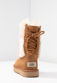 Brązowe zamszowe botki z kremowym futerkiem w środku, zawiązywane z tyłu i z gumową podeszwą. Widoczna metka z logo UGG po zewnętrznej stronie.
