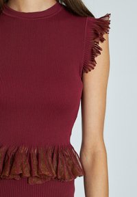Top senza maniche di colore bordeaux a coste con bordo trasparente e arricciato sulle spalle e in vita, indossato da una persona su uno sfondo neutro.