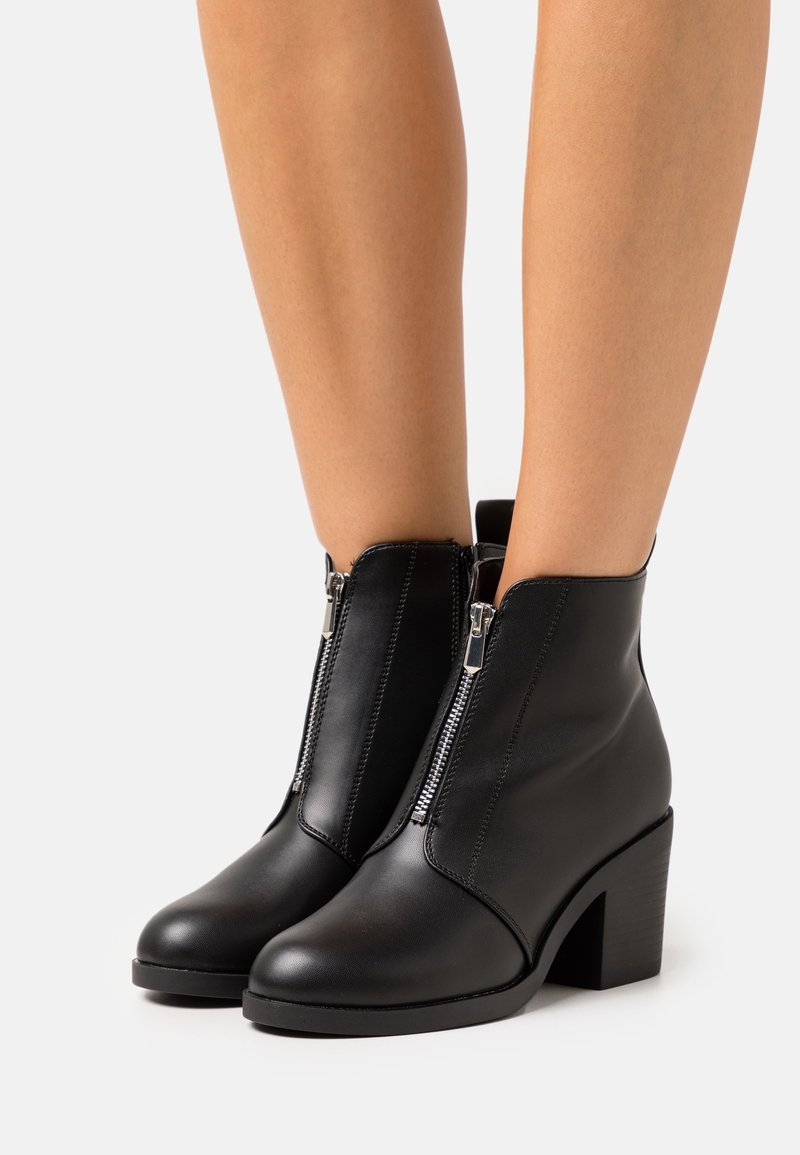Bottes cheville en cuir noir avec fermetures éclair argentées à l'avant et talons blocs portées sur des jambes nues sur fond blanc.