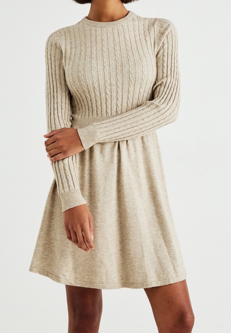 Robe en maille beige avec une texture côtelée, manches longues et corsage ajusté. Jupe évasée avec une forme discrète en A.