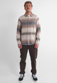 Camicia a righe con bottoni in tonalità beige, marrone e grigio; materiale in cotone; taschino sul petto; indossata sopra una t-shirt bianca e abbinata a pantaloni marroni.