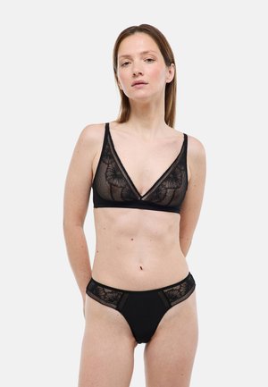 Femme portant un soutien-gorge en dentelle florale noire transparente et une culotte assortie, debout contre un fond blanc avec les mains derrière le dos.