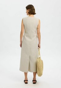 Ærmeløs, knælængde beige kjole med en lige silhuet og slits bagpå, parret med en vævet beige taske og brune sandaler.