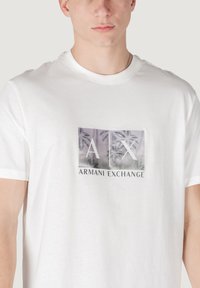 Camiseta de algodón blanco con un estampado gráfico que incluye "A|X" y "ARMANI EXCHANGE", complementada por una textura suave y un clásico cuello redondo.