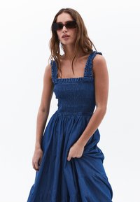 Denim-maksimekko, jossa on smockattu yläosa ja röyhelöhihnat. Syvän sininen väri, virtaviivainen kerroksellinen hame. Ei muita lisätietoja.