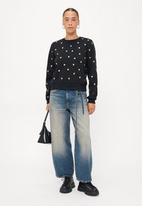 Sweatshirt noir avec des motifs en cœur dorés, associé à un jean large bleu délavé et des bottes noires épaisses. Sac à main noir tenu dans la main gauche.