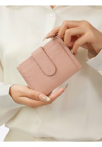 Portefeuille en cuir rose avec une finition texturée, doté d'un fermoir, de compartiments pour cartes et d'un compartiment zippé, tenu dans une main contre un fond clair.