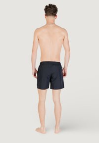 Pantaloni da bagno neri con vita elastica, lunghezza a metà coscia e texture del tessuto liscia. Persona è mostrata di spalle, a piedi nudi.