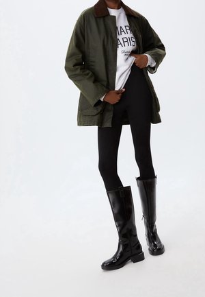 Personne portant une veste vert foncé, un sweat-shirt blanc imprimé, des leggings noirs et des bottes noires brillantes montant jusqu'aux genoux, debout devant un fond blanc.