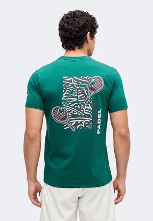 Άνδρας με την πλάτη φοράει πράσινο T-shirt με αφηρημένα γραφικά από ρακέτες παδέλ, μπάλες και δίχτυ και λευκά σορτς.
