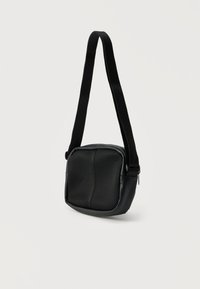 Μικρό μαύρο τσαντάκι ώμου με φαρδιά ρυθμιζόμενη λουρίδα και μπροστινή επιφάνεια με υφή από δίχτυ, κρεμασμένο μπροστά σε απλό φωτεινό φόντο.