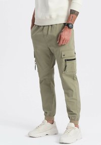 Mężczyzna w oliwkowych joggerach cargo z kieszeniami na zamek i guzikami, białych sneakersach, białej bluzie i czarnym zegarku na nadgarstku.