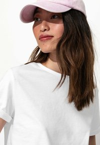 T-shirt en coton blanc avec des manches retroussées, associée à une casquette rose pâle. Les cheveux sont longs et ondulés, avec une texture naturelle.