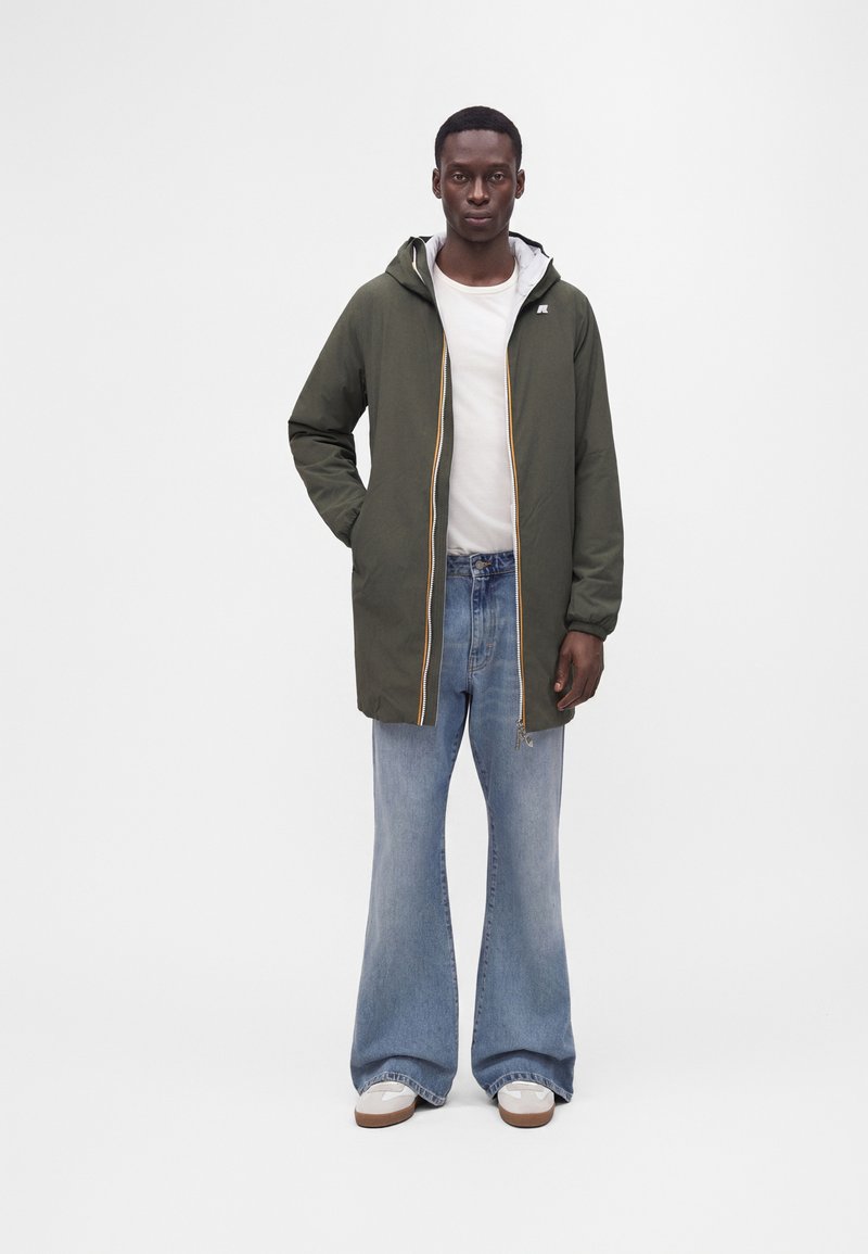 Άνδρας που φοράει ένα παλτό με κουκούλα σε χρώμα ελιάς πάνω από ένα λευκό μπλουζάκι, φαρδιά ανοιχτό μπλε τζιν και λευκά αθλητικά σε μονόχρωμο φόντο.
